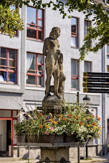 Figure on a fountain with flower frame in front of a building with windows, statue and monument in