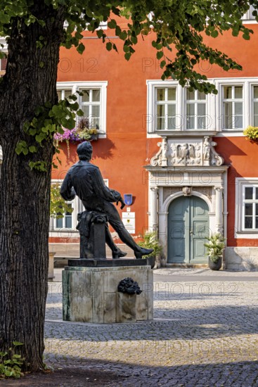 A statue in a square in front of a red, historic building with green door and blue windows, Johann