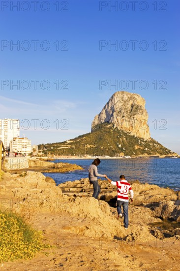 Two people on a rocky coast, natural park, in the background rock Penon de Ifach, landmark,