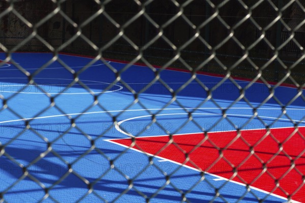Brightly colored basketball court features blue and red sections, seen through a chain-link fence.