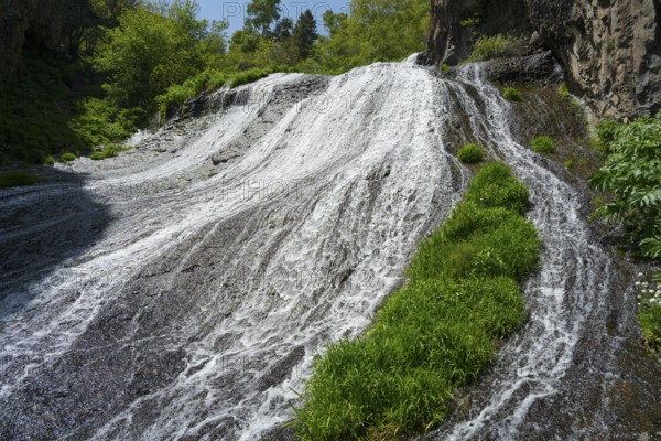 A wide waterfall flows steeply down, flanked by green plants and rocks in nature, Jermuk Waterfall,