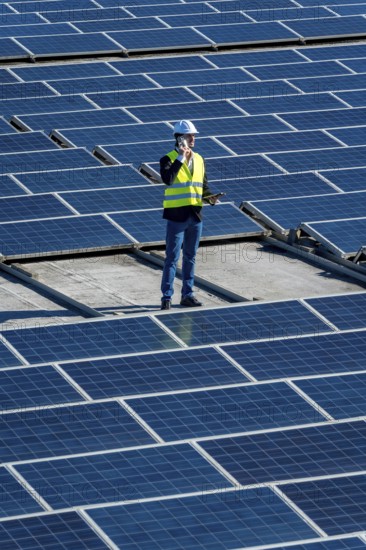 Engineer in hard hat and safety vest on commercial rooftop amid solar panels, using mobile phone