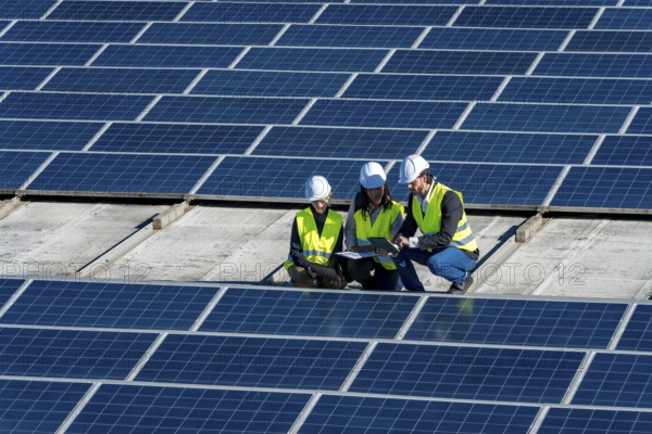 Team of engineers in hard hats and vests is discussing data on a tablet, working on a large solar