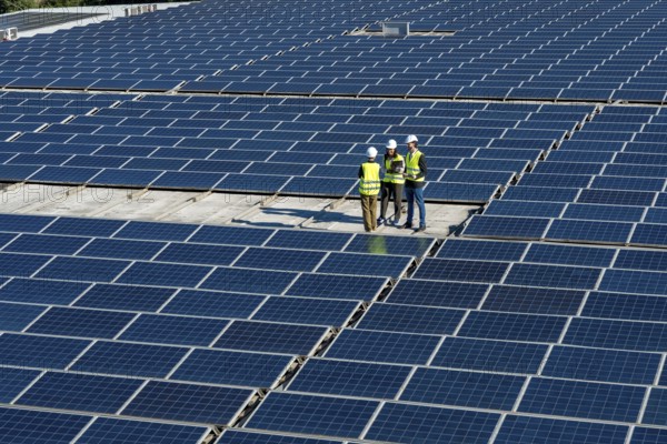 Engineers wearing safety vests and hard hats are standing on a large rooftop solar farm, reviewing