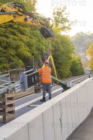 Excavator lifts objects on rails, construction worker wearing orange outfit, city in the