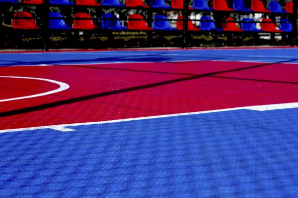 The basketball court features a striking red and blue design illuminated by bright sunlight. The