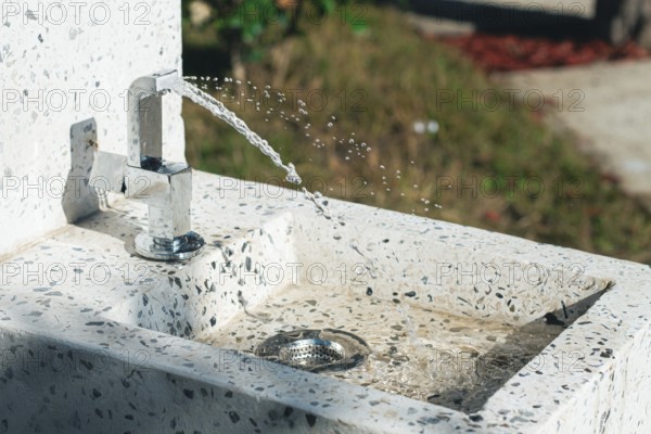 A clean tap fountain dispenses fresh water in a park surrounded by greenery. Sunlight shines on the