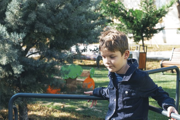 A young boy sits on a playground railing in a sunlit park, surrounded by greenery and trees. He