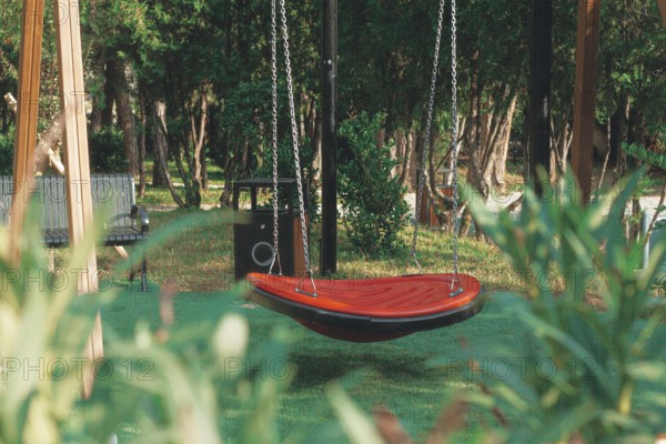 A vibrant red swing hangs suspended in a green park, surrounded by tall trees and wooden benches.