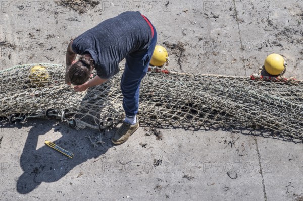 Seen from above, a fisherwoman in blue pants bends over a large fishing net spread out on the dry