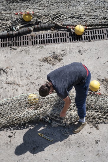 Seen from above, a fisherwoman in blue pants bends over a large fishing net spread out on the dry