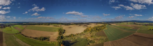 A large, open landscape shows green and brown fields with scattered trees. The sky is clear and