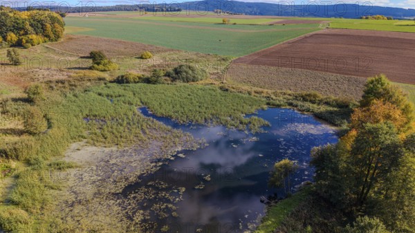 A quiet pond surrounded by meadows and trees. The colors of autumn are reflected while the sky is
