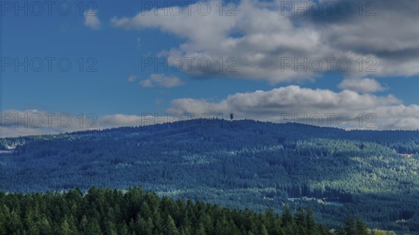 A green, wooded hill in the Bohemian Forest is visible under a blue sky with few clouds. Nature is