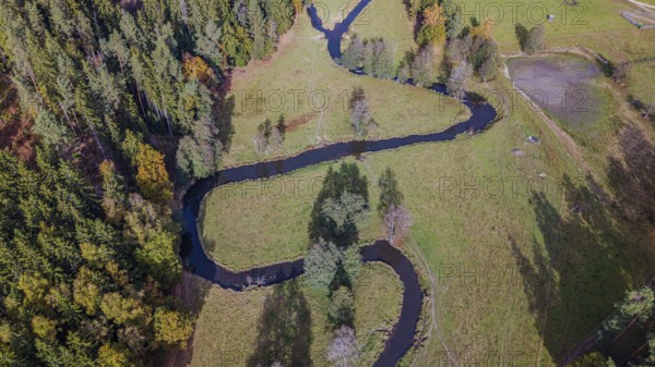 A clear river snakes through a green meadow, surrounded by colorful autumn trees. The picture shows