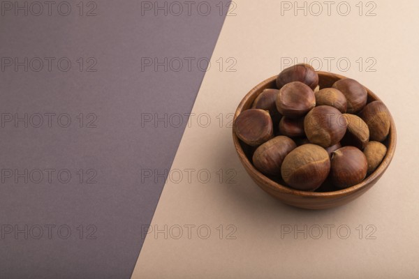 Wooden bowl with raw edible ?hestnuts on brown and gray paper pastel background, side view, copy