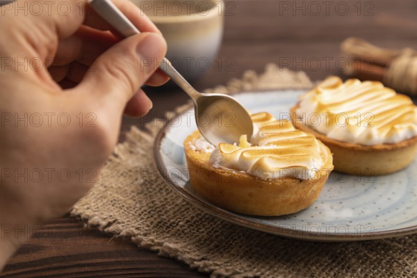 French lemon tart with meringue with hand on brown wooden background, cup of coffee, linen textile,