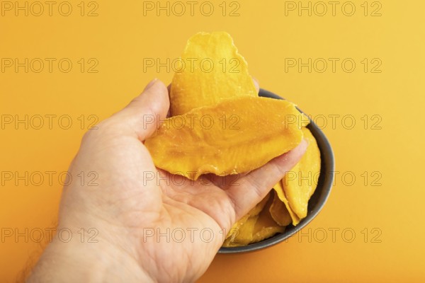 Dried Mango in blue ceramic bowl with hand on orange pastel paper background. Side view, close up.