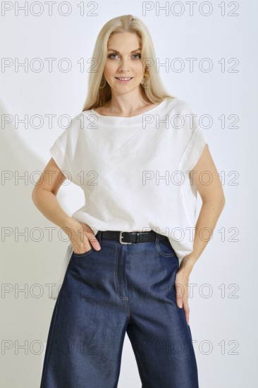 A blonde woman stands confidently in a bright studio, wearing a simple white top and loose blue