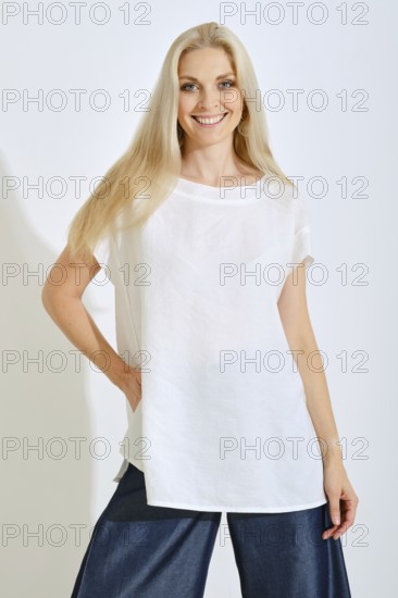 A woman stands smiling with long, light hair, dressed in a simple white top and dark denim pants.