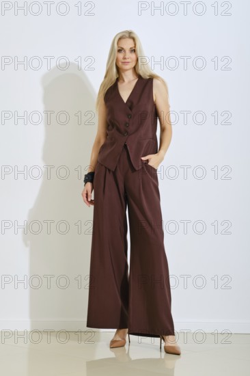 A woman stands elegantly in a chic brown tailored outfit that includes a fitted vest and wide-leg