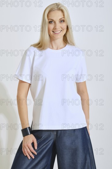 A woman stands confidently, wearing a plain white t-shirt paired with loose denim pants. She smiles