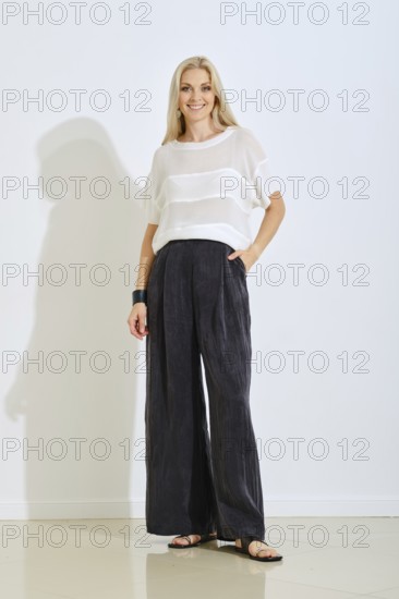 A model stands confidently indoors wearing a light top and loose black pants. The setting features