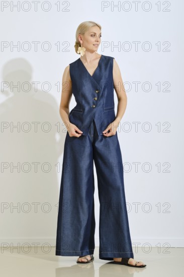 A woman stands gracefully in a stylish blue denim vest and wide-legged pants. She is indoors,