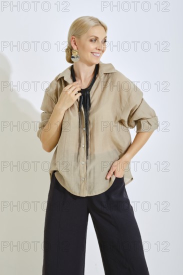 A woman is standing with a smile, dressed in a light beige shirt and dark pants. She wears a