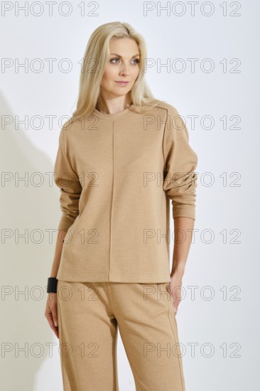 A confident woman stands elegantly in a bright studio, wearing a simple beige top and matching