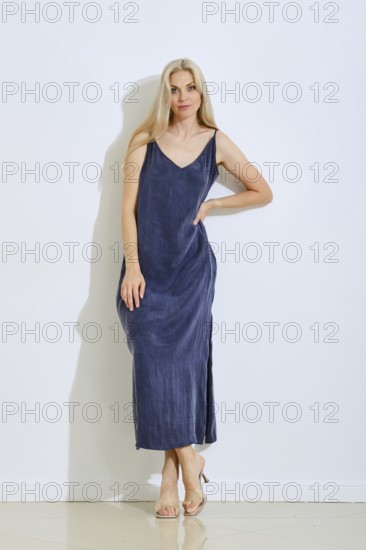 A woman stands gracefully against a plain white wall, wearing a long, sleeveless dress in a deep