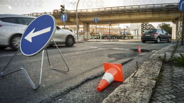 City street with large pothole marked by traffic cones and detour sign, cars passing by and