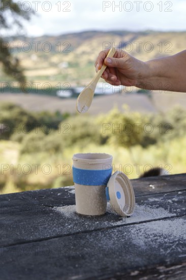 Person's hand adding a white powder with a wooden spoon into a reusable eco-friendly cup, enjoying