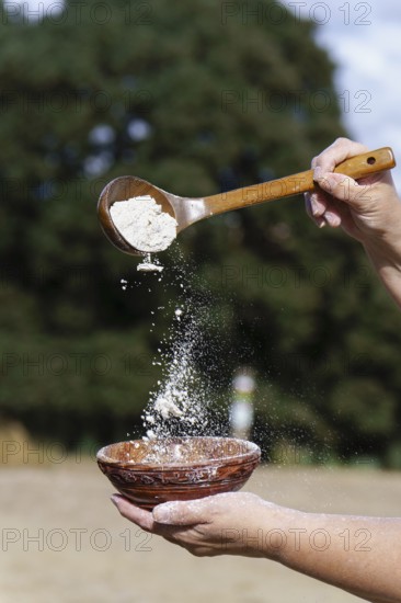 Hands sifting white flour from a wooden spoon into a rustic ceramic bowl, preparing ingredients for