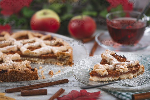 Sliced apple pie next to tea and apples, surrounded by autumn leaves and cinnamon sticks