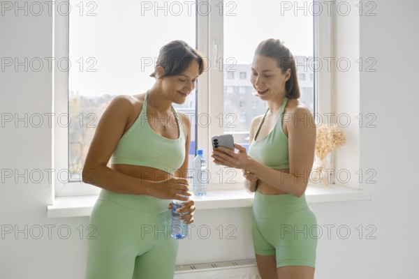 Two women in green workout outfits take a break during a fitness stretching class. They are smiling