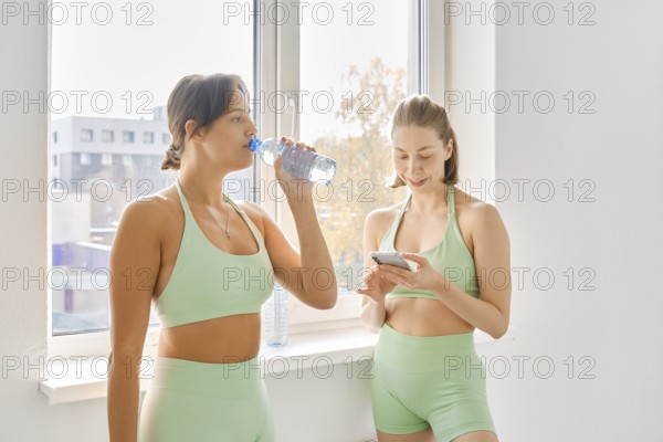 Two women take a break during a fitness stretching class in a bright indoor studio. One drinks