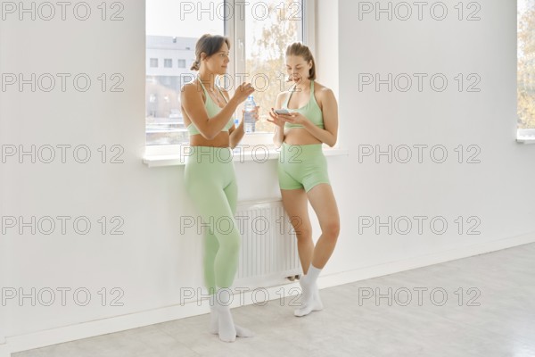 Two women in matching green workout outfits take a break during a fitness stretching class in a
