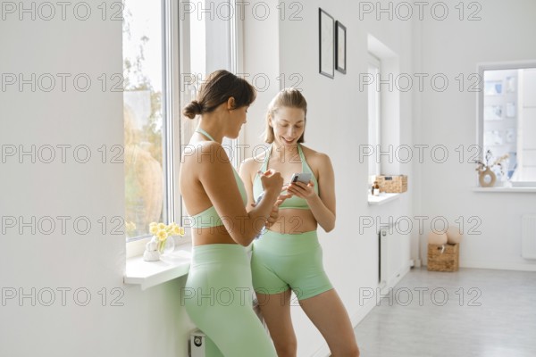 Two women in matching green workout outfits take a break during a fitness stretching class. They