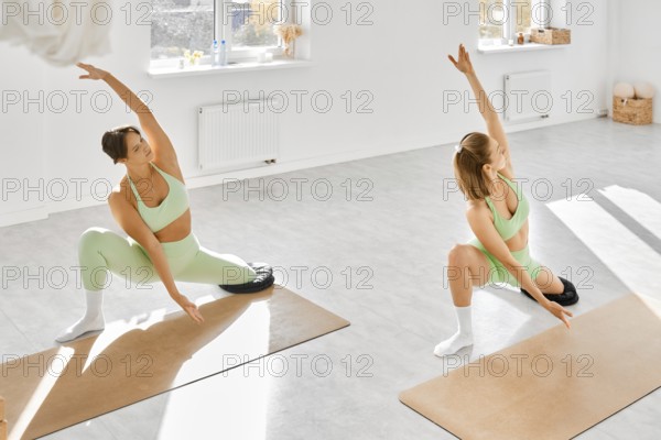 Two women are engaged in a yoga session in a well-lit studio filled with natural light. They
