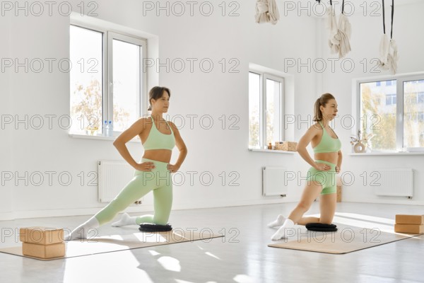 In a spacious studio filled with natural light, two women perform stretching exercises on mats.