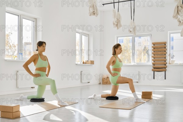 Two women practice lunges in a bright fitness studio. They use yoga mats and blocks for support.