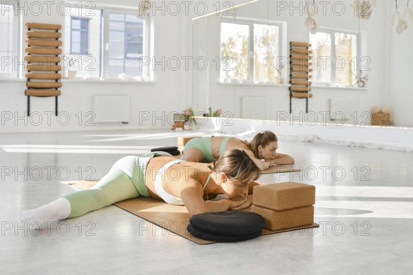 In a spacious and well-lit yoga studio, two women are engaged in a stretching routine on mats. They