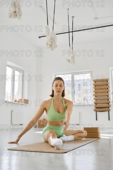 A young woman is seated on a yoga mat in a spacious, well-lit studio. She is stretching her body