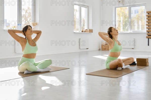 Two women are seated on yoga mats in a spacious, well-lit studio. They are dressed in matching