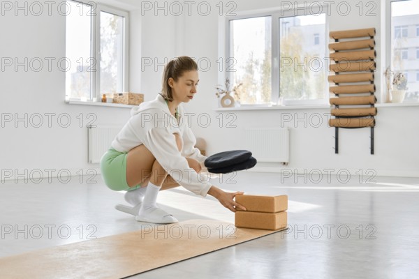 A woman squats on a yoga mat in a spacious studio. She is using stretching blocks to enhance her