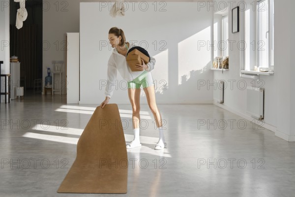 A young woman in light clothing is setting up a yoga mat in a spacious, sunlit studio. She is