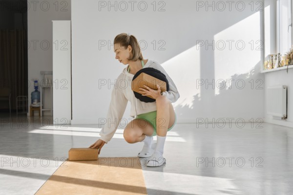 A woman in a white top and green shorts adjusts yoga props in a sunlit studio. The soft light