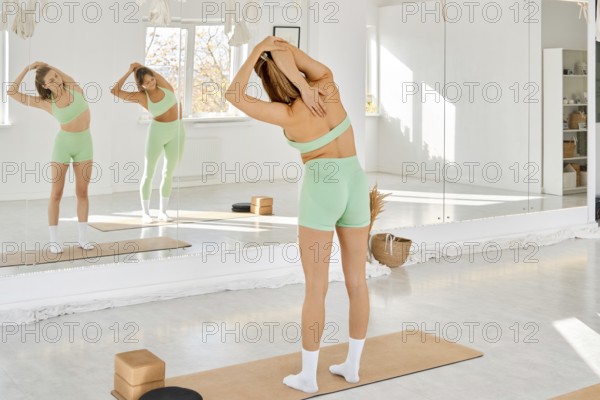 A woman performs a stretching exercise in a bright studio with large windows. She wears light green