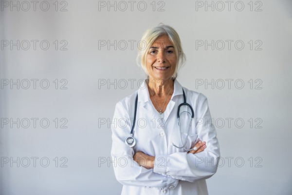 Senior female doctor with gray hair and a lab coat smiling confidently, standing against a neutral